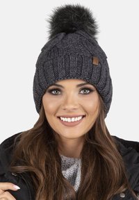 Bonnet gris tricoté avec une texture côtelée, surmonté d'un pompon en fausse fourrure noire. Comprend une petite étiquette de marque marron sur le côté.