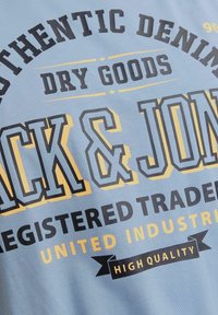 Camiseta de tela azul claro con texto en negrita impreso en negro y amarillo, incluyendo "Jack & Jones" y "Alta Calidad."
