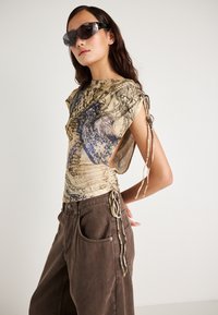 Korte mouwen top in beige met blauwe grafische ontwerpen, voorzien van zij-ruches en strikjes. Gecombineerd met loszittende bruine broeken.