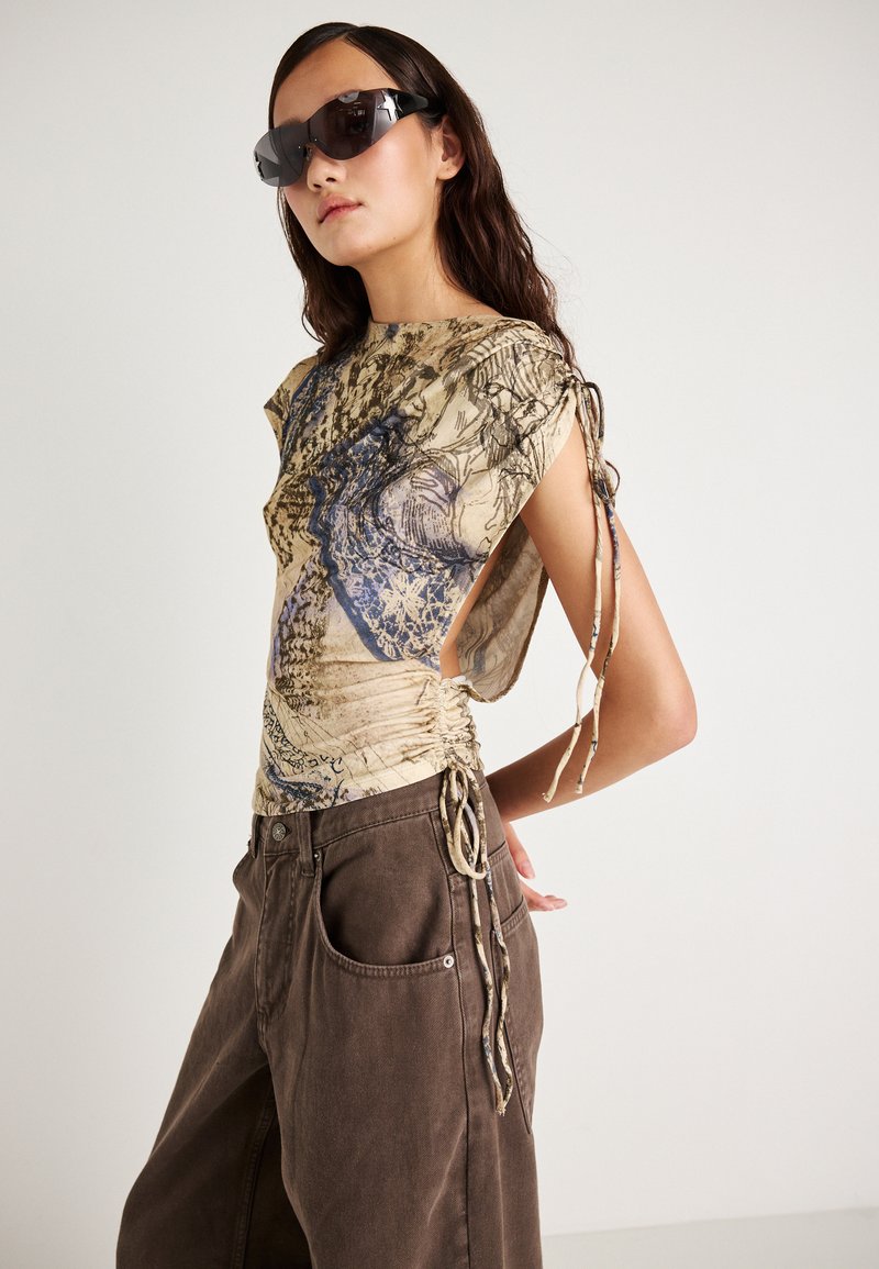 Korte mouwen top in beige met blauwe grafische ontwerpen, voorzien van zij-ruches en strikjes. Gecombineerd met loszittende bruine broeken.