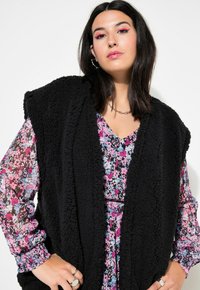 Schwarze Fleece-Weste mit strukturiertem Oberflächen, getragen über einem Blumenkleid mit pinken, blauen und schwarzen Mustern. Zubehör umfasst Ringe.