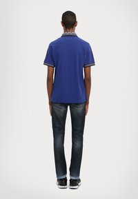 Modrá polo košeľa s vzorovaným golierom a manžetami, skombinovaná s tmavými denimovými džínsami. Oblečenie má neformálny dizajn a priliehavý strih.