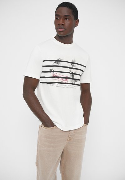 Λευκό βαμβακερό T-shirt με μαύρες οριζόντιες ρίγες και γραφικό σχέδιο. Λαιμόκοψη τύπου crew, χαλαρή εφαρμογή, συνδυασμένο με ανοιχτό μπεζ παντελόνια.