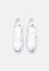 Ζευγάρι λευκών και μεταλλικά ασημί αθλητικών παπουτσιών με χοντρές λευκές κορδόνια, με την ένδειξη "Love Moschino" στα πλαϊνά και στις εσωτερικές σόλες, εμφανιζόμενα από πάνω.