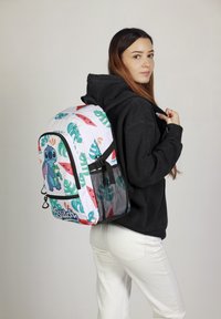 Mochila con una base blanca decorada con patrones de hojas tropicales verdes y un personaje de dibujos animados azul. Cintas negras, bolsillo lateral de malla y cierre con cremallera.