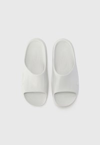 White slide sandals made of smooth rubber-like material, featuring an open toe and wide strap design with subtle branding on the side.