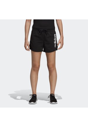 Osoba nosící černé kraťasy Adidas s bílým logem na levé noze, černé tenisky, stojící na jednobarevném světlém pozadí.