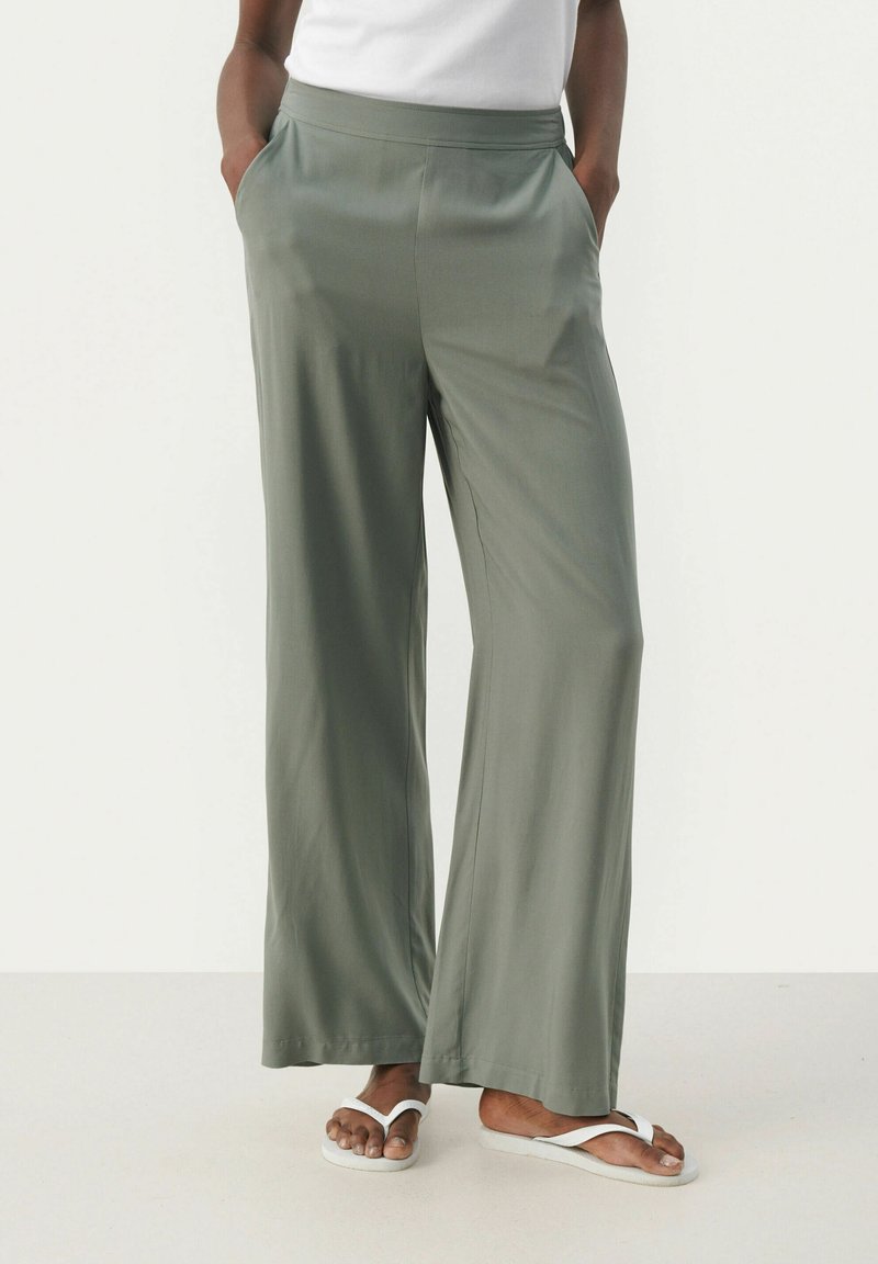 Pantalons larges en vert olive, fabriqués dans un tissu lisse. Dotés d'une taille élastique et de poches latérales. Portés avec des tongs blanches.