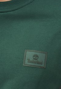 Πράσινο πλεκτό ύφασμα με ραμμένο ορθογώνιο έμπλαστρο που φέρει το λογότυπο δέντρου Timberland και το όνομα της μάρκας στο ίδιο πράσινο χρώμα.