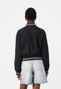 Chaqueta bomber negra con rayas blancas en el cuello y los puños. Tela ligera y cintura elástica. Combinada con pantalones cortos de mezclilla claros.