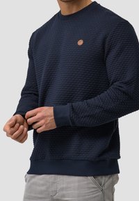 Marineblauwe sweatstof met structuur, voorzien van een ronde halslijn en geribbelde manchetten. Heeft een klein bruin merkpatch op de borst.