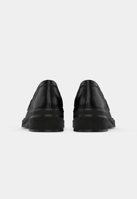 Paire de chaussures habillées en cuir noir à talons épais vues de dos sur un fond uni gris clair.