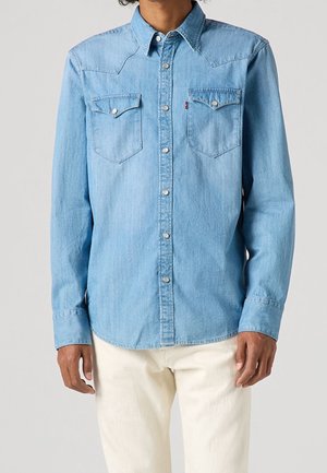 Persona que lleva una camisa vaquera azul claro con botones y bolsillos en el pecho, y pantalones blancos, mostrada desde la mitad del torso hasta las caderas.