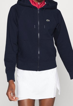 Person iført navy lynlås-hættetrøje med forlommer og logo, rød top under og hvid nederdel med sideslids.
