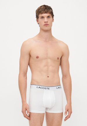 Jeune homme aux cheveux bruns courts portant des boxers blancs Lacoste sur un fond clair.