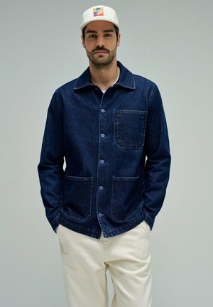 Hombre con gorra blanca con parche colorido, chaqueta vaquera azul oscuro con botones y tres bolsillos, y pantalones beige claro, de pie con las manos en los bolsillos.