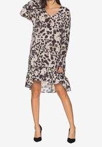 Kvinna iklädd långärmad klänning med leopardtryck, volangfåll och svarta högklackade skor, poserar med en hand på huvudet.