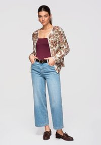 Femme debout les mains dans les poches, portant une veste à motifs floraux, un haut bordeaux, un jean bleu clair, une ceinture noire et des mocassins marron.