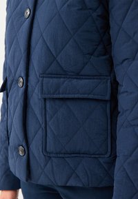 Giacca trapuntata blu navy con cuciture a rombo, chiusura con bottoni e tasca a patta. Il tessuto sembra morbido e leggermente texturizzato.