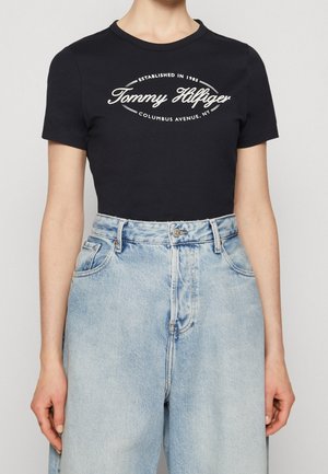 Czarna dopasowana koszulka z białym napisem "Tommy Hilfiger" oraz jasnoniebieskie dżinsy z wysokim stanem, ręce swobodnie opuszczone wzdłuż ciała.