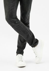Weiße Sneaker mit Schnürsenkeln, gepolsterten Zungen und strukturierten Sohlen, kombiniert mit schwarzen Slim-Fit-Jeans, die einen ausgebleichten Look und feine Abriebe aufweisen.