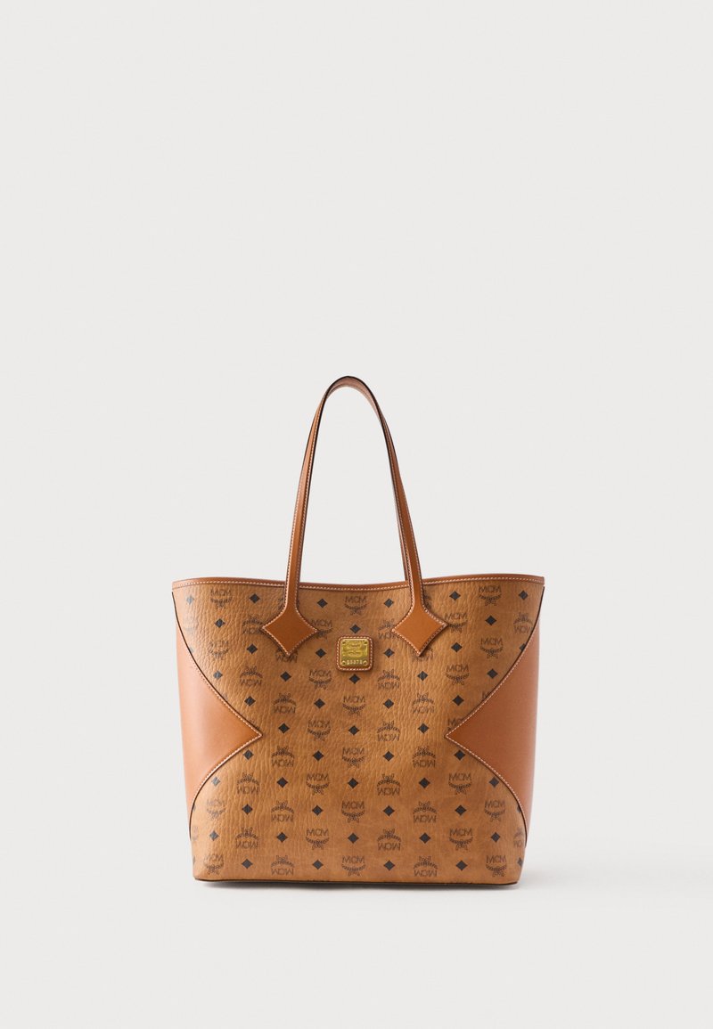 Καφέ δερμάτινη τσάντα ώμου με μοτίβο λογότυπου MCM, δύο χειρολαβές και χρυσή τετράγωνη πινακίδα λογότυπου σε απλό ανοιχτό φόντο.