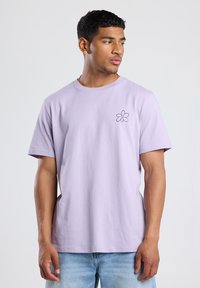 Laventelin värinen puuvillainen t-paita, jossa on lyhyet hihat, pyöreä kaula-aukko ja musta kukka brodeerattu design rinnassa. Rentoa istuvuutta, sileä pinta.