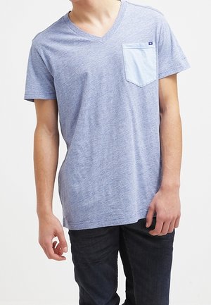 Junger Mann, der ein hellblaues V-Ausschnitt-T-Shirt mit kontrastierender Brusttasche und dunkle Jeans trägt, vor einem schlichten hellen Hintergrund stehend.