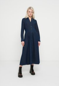 Mørk denimskjortekjole med lange ærmer, knaplukning foran og A-formet skæring. Båret med sorte snørestøvler med tykke såler.