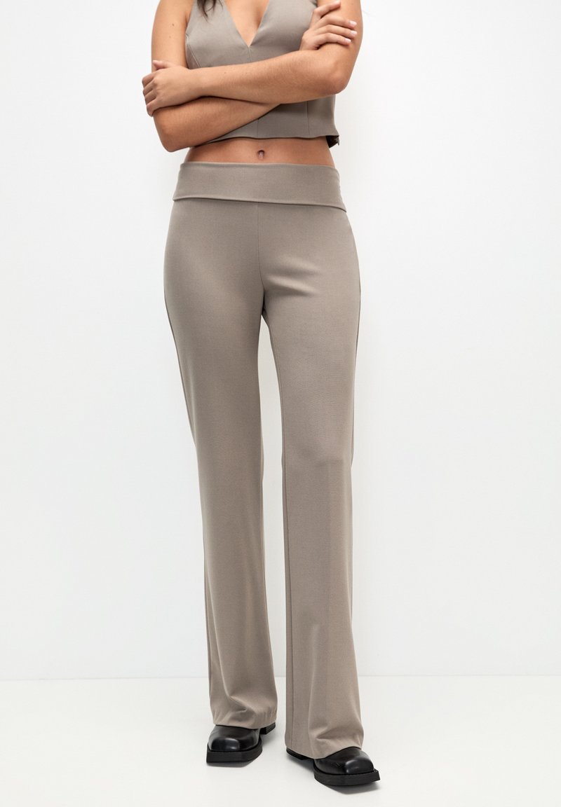 Frau trägt taupefarbene, hochgeschnittene, weit geschnittene Hose und ein passendes, kurzes Oberteil mit verschränkten Armen und steht vor einem schlichten weißen Hintergrund.