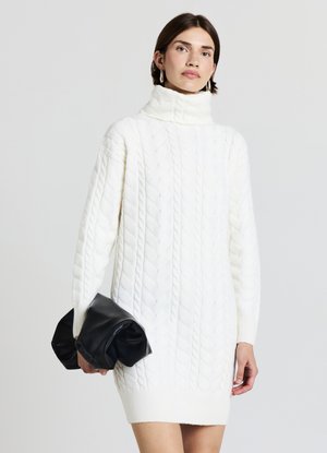 Robe-pull en maille torsadée blanche avec col montant, ourlet côtelé et manches longues. Associée à une pochette noire froissée.