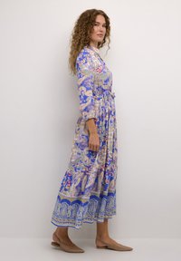 Maksi obleka v modri barvi z vzorcem paisley, dolgi rokavi, zategnjen pas in razširjen rob. Oblečena z bež natikači.