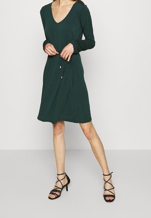Kobieta ubrana w ciemnozieloną, długorękawową sukienkę sięgającą do kolan z dekoltem w kształcie litery V, do której dobrała czarne sandały na wysokim obcasie z paskami.