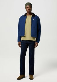 Chaqueta de lona azul marina con cremallera, camisa verde oliva debajo, pantalones vaqueros rectos azul oscuro y botas marrones texturizadas. Gorro de punto con acento de logo.