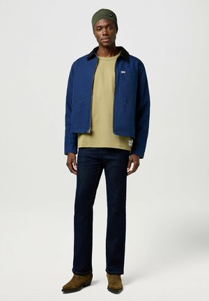Chaqueta de lona azul marina con cremallera, camisa verde oliva debajo, pantalones vaqueros rectos azul oscuro y botas marrones texturizadas. Gorro de punto con acento de logo.