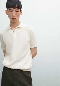 Polo shirt a costine crema con maniche corte, colletto a contrasto e bottoni a tre vie, abbinato a pantaloni scuri. Texture liscia.
