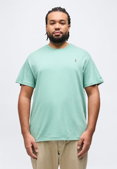 Homme avec cheveux tressés et barbe portant un t-shirt à manches courtes vert menthe et un pantalon beige, debout devant un fond clair uni.