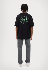 Black cotton t-shirt with green "RESORT CLUB" text and stars graphic on the back, paired with gray denim pants and white sneakers.