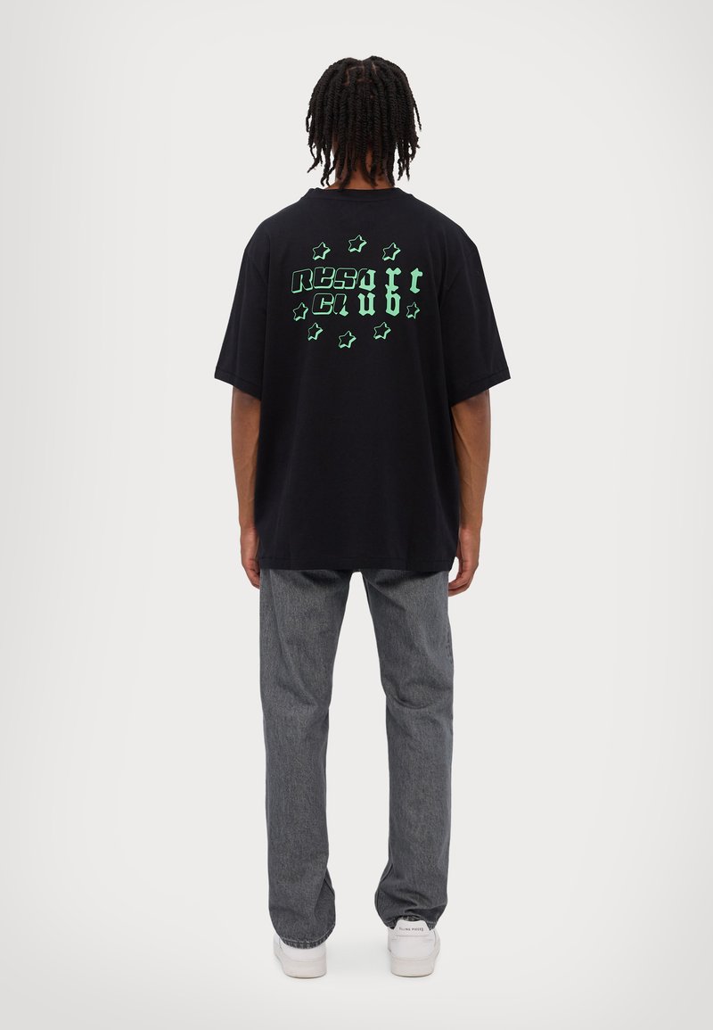 Black cotton t-shirt with green "RESORT CLUB" text and stars graphic on the back, paired with gray denim pants and white sneakers.