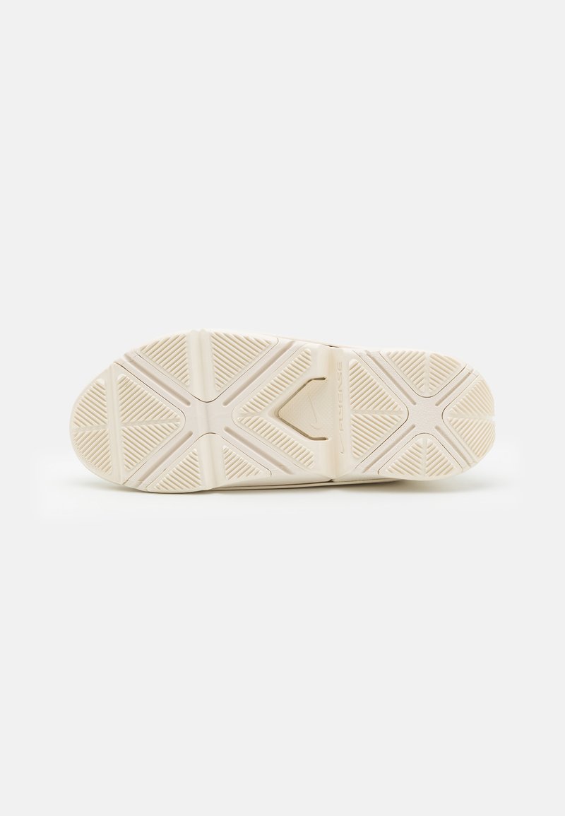 Semelle de sneaker en caoutchouc blanc présentant un motif de grip texturé et un détail de logo central. Le design inclut des rainures pour la traction.