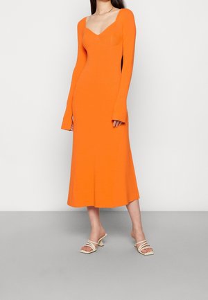 Kvinde iført en lang, tætsiddende orange kjole med lange ærmer og hvide sandaler med hæl og stropper, stående på en grå gulv mod en hvid væg.