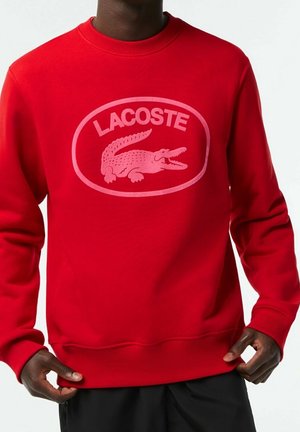 Mężczyzna w jasnoczerwonej bluzie Lacoste z różowym logo krokodyla i napisem, poprawiający dół bluzy, na jednolitym białym tle.