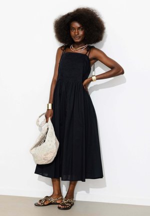 Femme aux grands cheveux bouclés portant une robe noire sans manches, des bracelets en or, des colliers superposés, des sandales à imprimé léopard et tenant un sac à main beige à motifs.