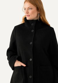 Cappotto in misto lana nera con colletto alto, bottoni frontali e ampie tasche laterali. Tessuto morbido e vestibilità rilassata.