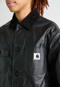 Giacca con colletto in corduroy nero realizzata in tessuto lucido, con due tasche sul petto e il logo Carhartt sulla tasca sinistra.