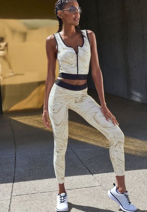 Athletic set in cream with gold marbling, featuring a zip-up sleeveless top and high-waisted leggings; paired with white sneakers.