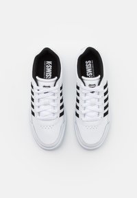 Biele športové topánky s čiernymi pruhmi, šnurovací dizajn, hladký kožený materiál a polstrovaný golier pre pohodlie. Logo je viditeľné na jazyku.