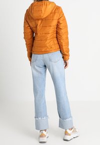 Orange pufferjacka med huva, med kviltad design. Matchad med ljusblå vida jeans med fransade fållar och vita sneakers med detaljer.