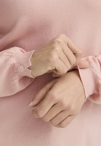 Pull tricoté rose clair avec poignets côtelés, orné d'un détail à boutons. Gros plan sur des mains ajustant le poignet. Texture douce visible.