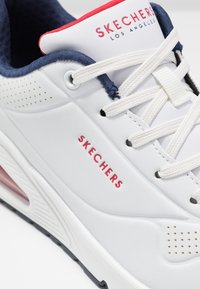 Bílé tenisky vyrobené z kůže s texturovaným modrým a červeným polstrovaným límcem. Mají perforace a výrazné logo "SKECHERS" v červené barvě.